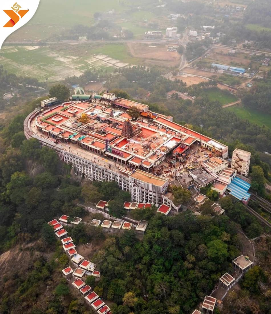 An aerial view of the Palani Murugan Temple situated on a hill in Tamil Nadu, featuring a sprawling structure with colorful roofs and lush greenery surrounding it, alongside pathways leading to the temple.